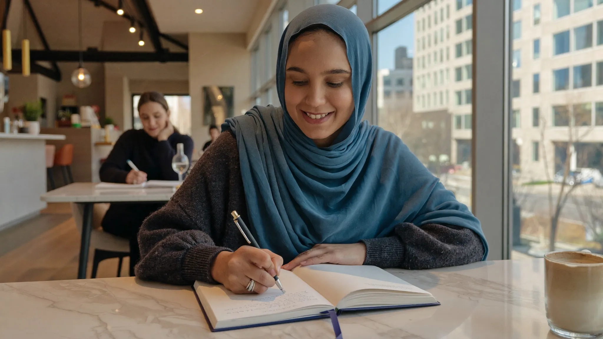 Humna Raza writing in a journal at a sunlit cafe table.